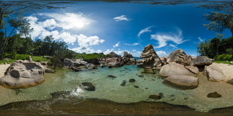 Obraz premium Shallow lagoon with crystal-clear water surrounded by smooth, large boulders and vibrant greenery under a bright sun. La Digue, Seychelles. 360 Degree view.