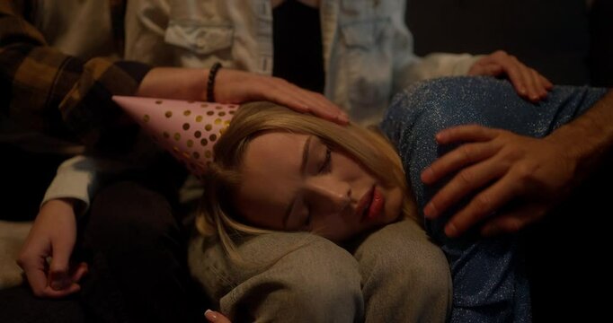 A sleepy and sad girl in a festive cap lies on the laps of her friends and they support and stroke her at home in the evening