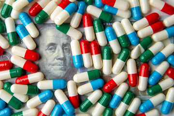 A close-up image of colorful capsules scattered on a US dollar bill. The high cost of medication...