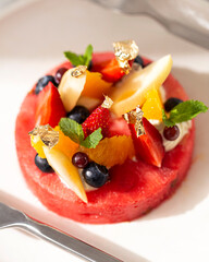 Close-up of a fresh watermelon fruit pizza topped with berries, citrus, pear, cream, mint, and edible gold leaf.
