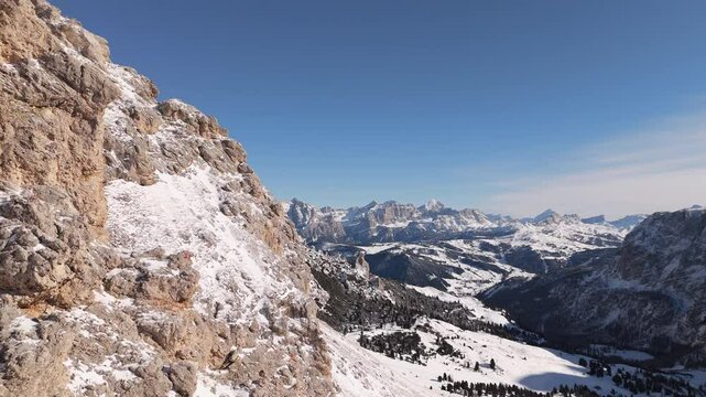 Snow-covered cliff face overlooks a vast winter landscape of the Dolomites, with forested valleys and distant mountain peaks stretching to the horizon.