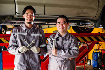 Two skilled Asian auto mechanics in gray uniforms stand confidently in garage, holding wrenches. Background with lifted car, workshop tool equipments, symbol of teamwork, expertise, car repair service