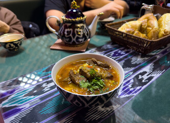 A bowl of soup sits on a table with a tea set and a basket of bread