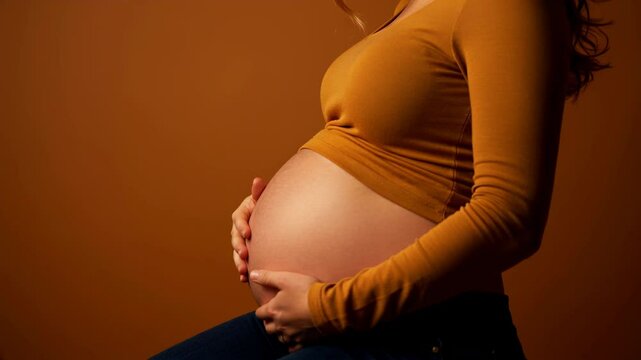 Pregnant woman in mustard sweater gently touching her belly against warm brown background. Motherhood and expectancy concept. pregnancy clinics, maternity wear, family blogs. Horizontal video