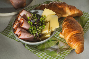 early morning breakfast. ham, cheese and sprouts in bowl, fresh croissants on cheerful green cloth, sunrays and shadow high angle view