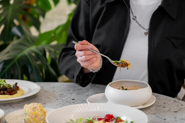 A close-up of a person's hand holding a fork, poised to take a bite of food from a bowl. In the background, a beautifully arranged table with various dishes and green foliage.