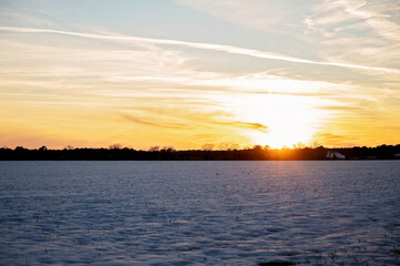 Fototapeta premium A snow-covered field at sunset with a golden sky and distant farm