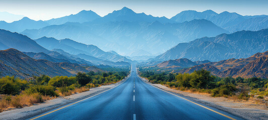 Naklejka premium Scenic open road stretches into distance, surrounded by majestic mountains and lush greenery under clear blue sky.