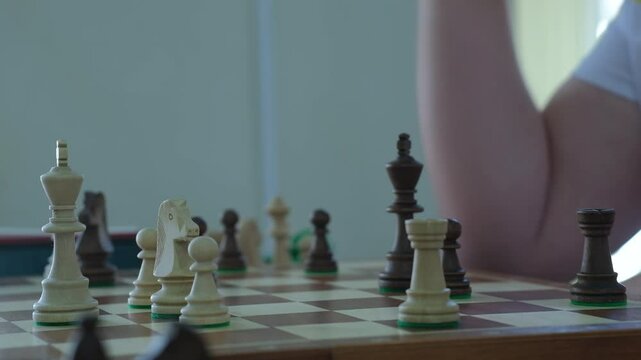Player takes King in hand to make move sitting at table. Person in T-shirt stops time after moving piece. Competitive game of chess with timer