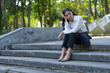 Thoughtful young woman in business attire sitting on concrete steps in a park, surrounded by lush...