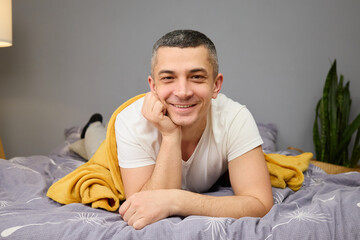 A man in a white t-shirt lounges comfortably on a bed draped with a yellow blanket. He smiles...
