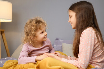 Two girls, one with curly hair and the other with straight hair, sit closely together on a bed,...
