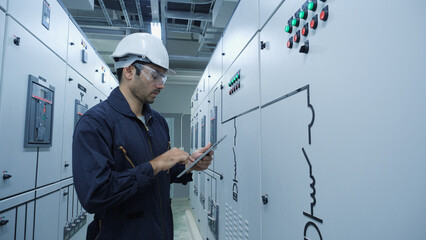 Industrial engineer wearing helmet and protective glasses using tablet to inspect electrical control panel in factory. System performance, troubleshooting and automation in power distribution.