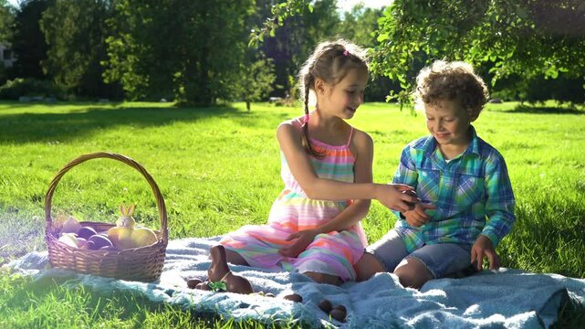 Children enjoying Easter picnic in a sunny park, eating chocolate eggs. A wicker basket with decorated eggs and a chocolate bunny sits on a blanket. Green grass and trees create a beautiful spring