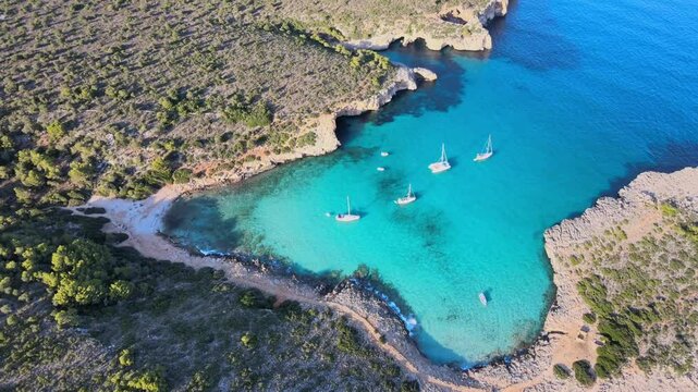 Discover the tranquil beauty of Cala Varques in Majorca, where crystal clear waters meet stunning coastal landscapes. Enjoy the sight of sailing boats anchored in a serene cove surrounded by nature.
