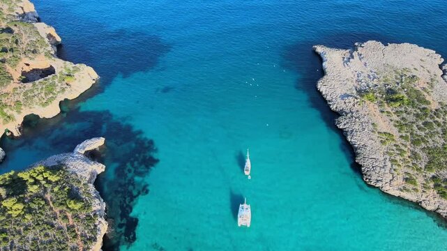 Discover the breathtaking beauty of Cala Varques in Porto Cristo, Majorca, where crystal-clear waters meet rugged cliffs. Enjoy a tranquil day with boats dotting the shoreline.