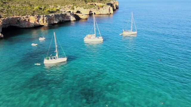 Sailboats gently float in the turquoise waters of Cala Varques, nestled near Porto Cristo in Majorca. This beautiful cove features stunning cliffs and serene surroundings, perfect for relaxation.