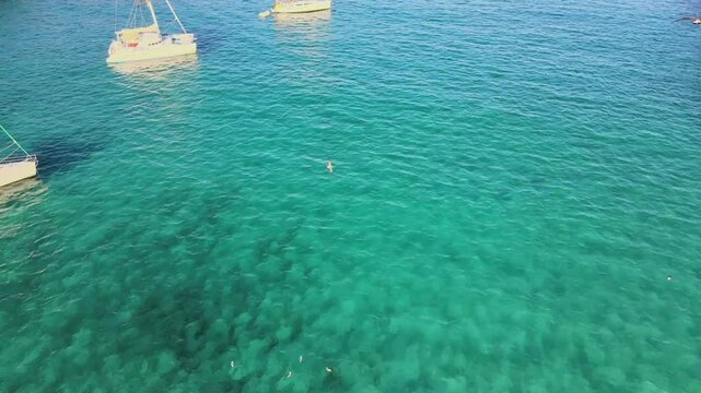 Experience the beauty of Cala Varques in Porto Cristo, Majorca. Sailboats glide over clear turquoise waters, inviting swimmers to enjoy a refreshing dip in this serene paradise.