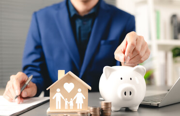 Family saving money concept. Businessman deposits coin into piggy bank with wooden house model,...