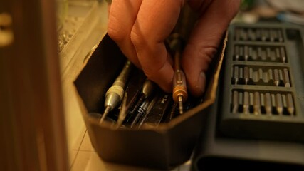 Close-up hand of unrecognizable repairman carefully selecting precision screwdriver from box containing various small tools. Concept of meticulous tool organization in workshop or repair setting.