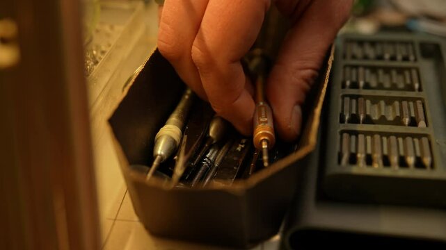 Close-up hand of unrecognizable repairman carefully selecting precision screwdriver from box containing various small tools. Concept of meticulous tool organization in workshop or repair setting.
