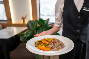 A waiter is serving a plate featuring golden-brown fried patties, pickles, and a side of creamy mushroom sauce. The background features a cozy, modern restaurant setting.