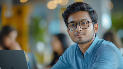 Obraz premium Focused Young Man in Blue Shirt Working on Laptop – Modern Office Environment with Professionals – Serious & Productive Business Atmosphere in a Collaborative Workspace with Large Windows