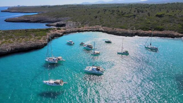 Experience a serene day on the water at Cala Varques, Majorca. Sail alongside picturesque boats in crystal clear turquoise waters surrounded by lush greenery and rocky cliffs.