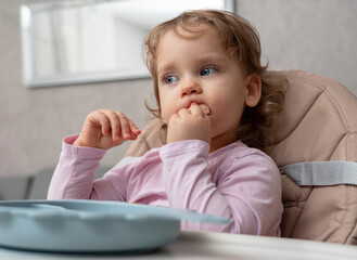 Fototapeta premium A thoughtful toddler focused on self-feeding while chewing a piece of sausage.