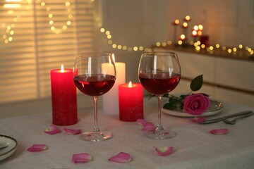 Romantic place setting with red wine, burning candles, rose and petals on table against blurred...
