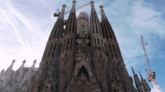 Upward panning shot of Sagrada Familia, showcasing intricate facades and towering spires. Sunlight shines through the architecture.