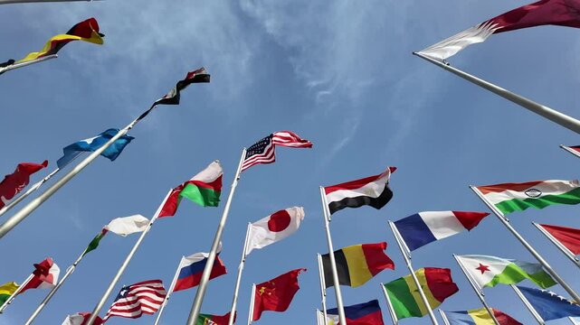 A collection of international flags waving on tall poles against a bright blue sky, symbolizing global unity and diversity.
