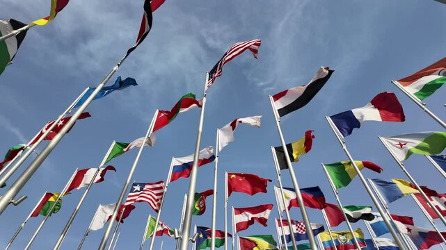 A collection of international flags waving on tall poles against a bright blue sky, symbolizing global unity and diversity.