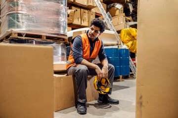 Tired interracial warehouse worker with helmet in hands sitting on box surrounded by other boxes.