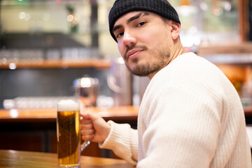 A young Latin man in a beanie holding a pint of beer in a cozy pub. Warm, friendly atmosphere in a European bar setting