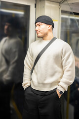 A young man in a white sweater standing inside a subway train, reflecting on the window