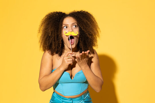 Playful woman gesturing with mustache prop against yellow background