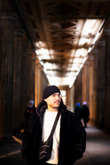 A young man in winter fashion standing in a grand, historic corridor with glowing lights and...