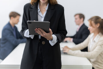 Professional woman stands confidently with tablet in a modern office while colleagues work on laptops in the background