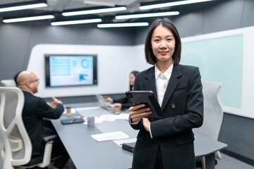 Businesswoman standing poses, group meeting around a table in modern office, documents, laptops and graphs are placed, everyone brainstorming, planning sales, digital marketing and growth strategies.
