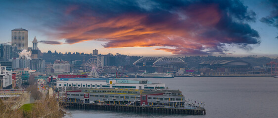 Aerial view of Seattle, Washington, featuring the Great Wheel, Smith Tower, Lumen Field, T Mobile Park, a pier, a cargo ship, and a vibrant sky. © Aerial Film Studio