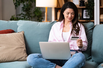 Young smiling happy independent woman sitting at home on sofa, making purchase. Online shopping from home on laptop computer with bank credit card.