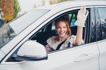Cheerful young woman in vintage attire waves from a white car on a sunny day