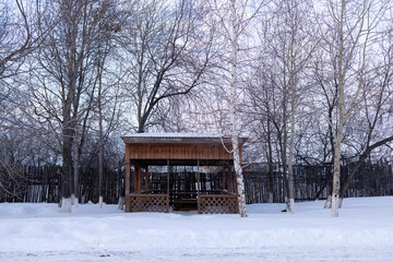 A wooden gazebo stands in snowdrifts by the road. Bare birch trees nearby.