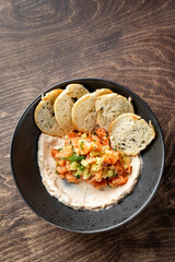 A close-up of a bowl featuring a creamy dip topped with fresh diced vegetables and herbs, accompanied by crispy toasted bread slices. Perfect for appetizers or gourmet dining.