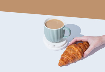 Hand holding fresh croissant on white broun background and cup of coffee with milk.