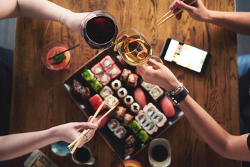 Friends gather around a wooden table, enjoying sushi and drinks. Hands reach for fresh sushi rolls, creating a cozy and social dining atmosphere