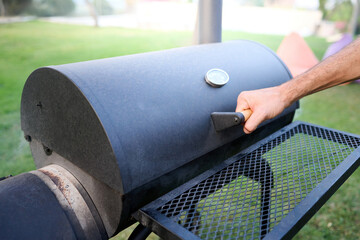 Unrecognizable man grabs handle to open lid with thermometer of smoker. Cooking on outdoor barbecue in backyard at home.