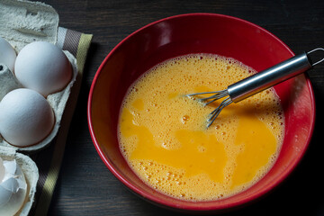 Whisking eggs. Whipped raw eggs in a red bowl with whisk for omelet , closeup. Breakfast preparation, rustic style