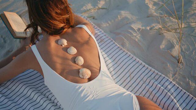 Woman body lying beach with seashells on back closeup. Lady reading novel